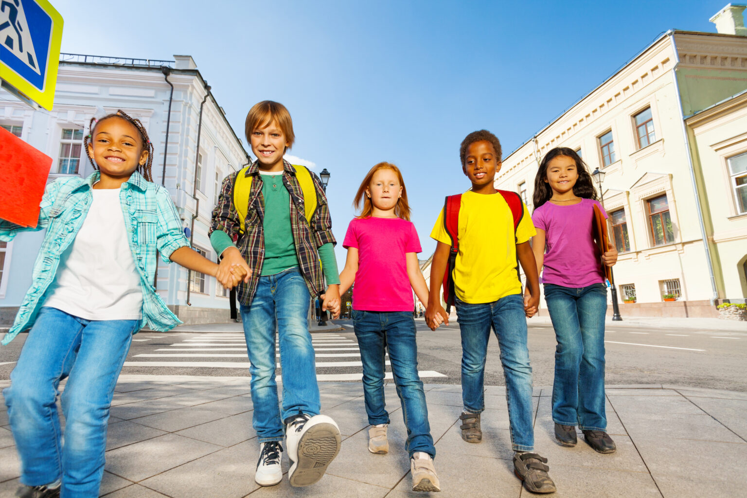 International children wear bags, walk in row together and hold hands of each other on the city street in summer time