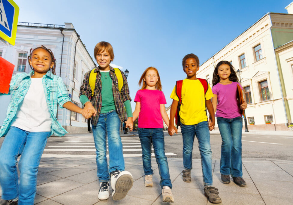 International children wear bags, walk in row together and hold hands of each other on the city street in summer time